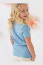 A young blonde girl wearing a light blue short-sleeve t-shirt and cream pants, shown from a side-back angle against a bright white background with soft pink decorative elements. 