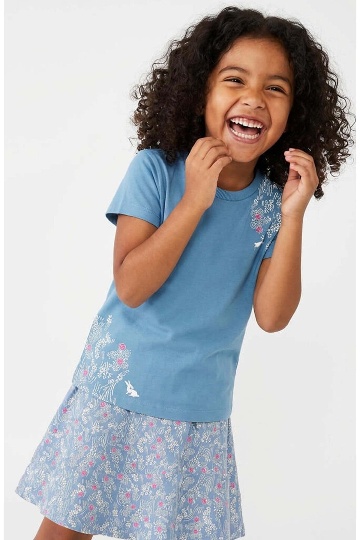 A young girl with curly dark hair is smiling and laughing while wearing a light blue short-sleeve t-shirt with a floral patterned skort, photographed against a clean white studio background.