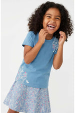 A young girl with curly dark hair is smiling and laughing while wearing a light blue short-sleeve t-shirt with a floral patterned skort, photographed against a clean white studio background.