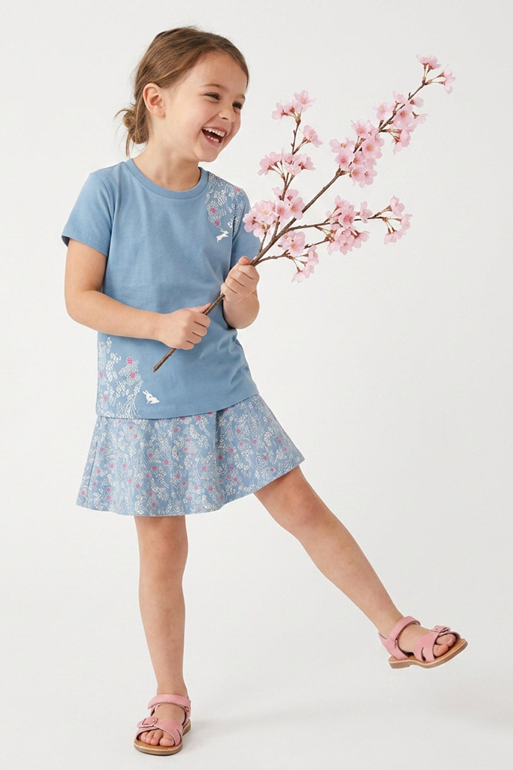 A young girl wearing a light blue short-sleeve dress with a floral pattern and butterfly details, holding a pink cherry blossom branch against a clean white studio background.