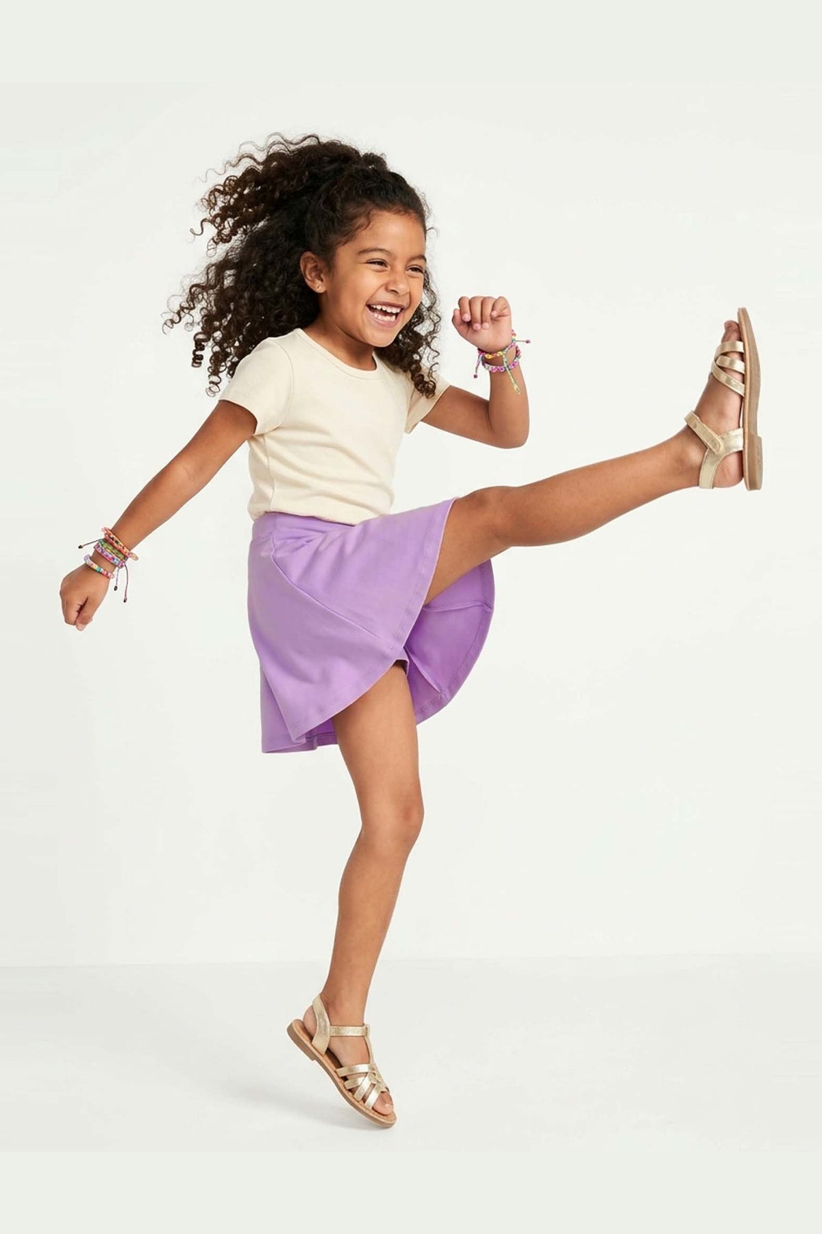 Child wearing a white top and purple skirt with gold sandals on a white background 