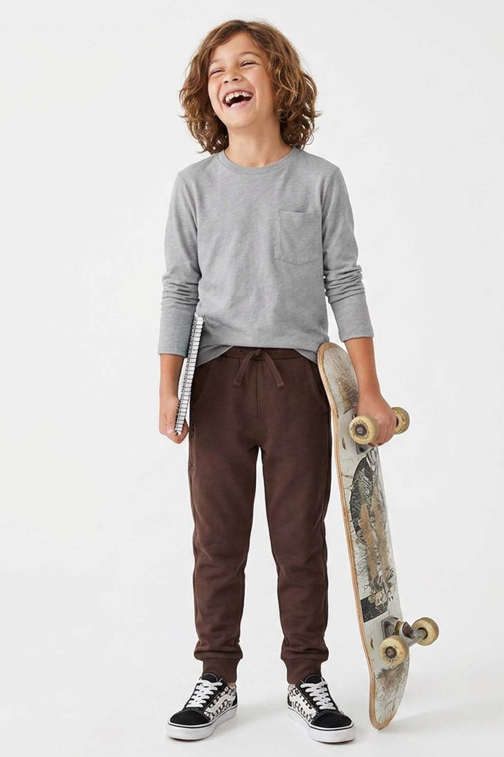 A young boy with curly brown hair stands smiling and laughing while holding a skateboard. He is wearing a light gray long-sleeve crewneck shirt, dark brown drawstring sweatpants, and black and white sneakers, set against a plain white studio background. 