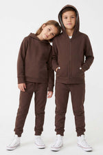 A child's matching two-piece tracksuit in dark brown, shown on two models (a boy and a girl) against a plain white studio background. The set includes a zip-up hoodie with a front pocket and matching jogger pants. 