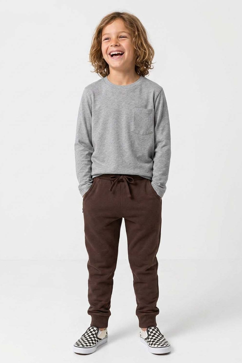 A young boy with curly brown hair stands smiling in a studio setting, wearing a heather gray long-sleeve crewneck sweatshirt, dark brown jogger pants, and black-and-white checkered slip-on shoes. 