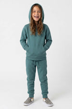 A young girl smiling while wearing a matching teal green hooded sweatshirt and jogger pants set, paired with slip-on shoes, against a plain light gray studio background. 