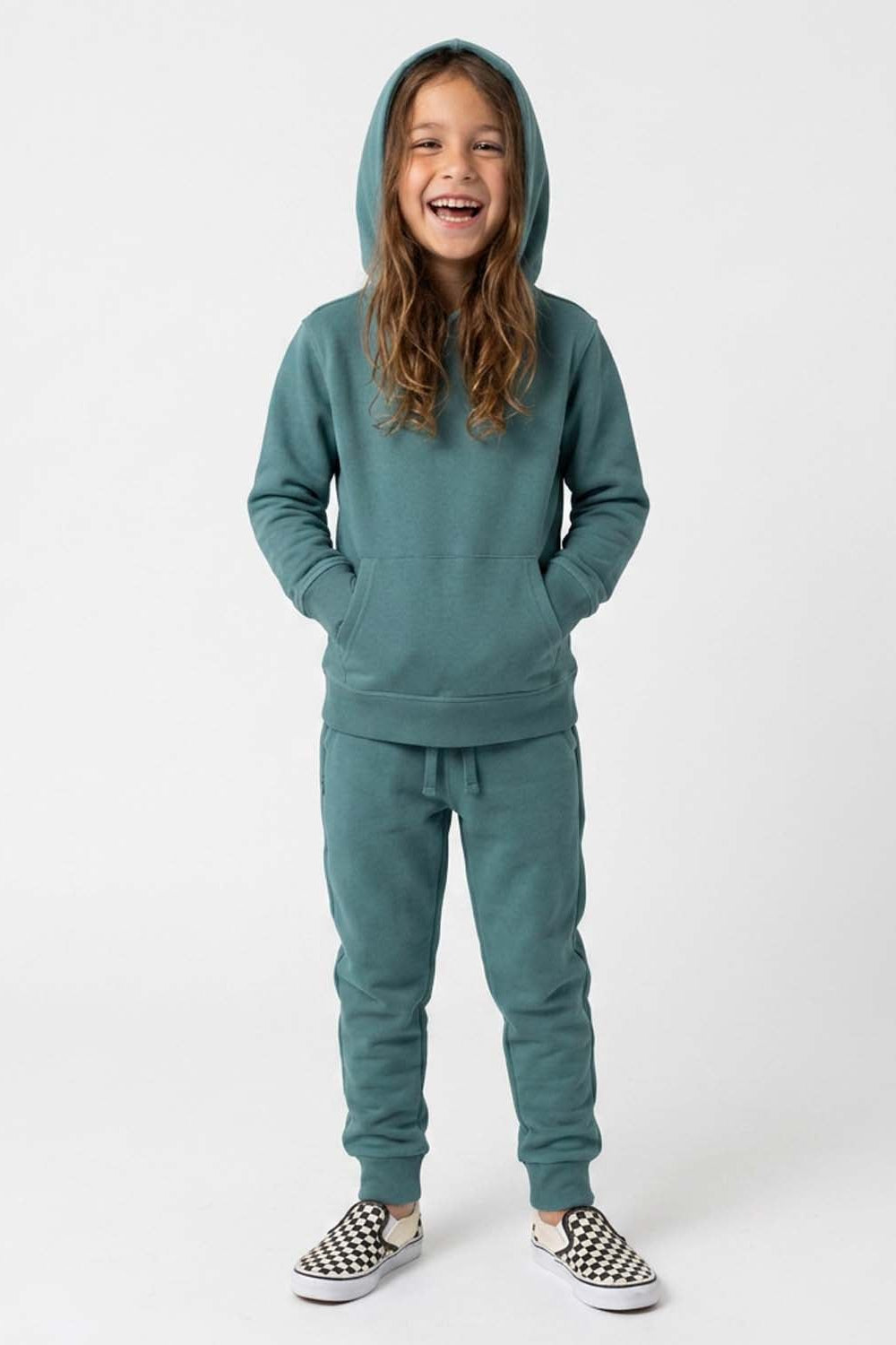 A young girl smiling while wearing a matching teal green hooded sweatshirt and jogger pants set, paired with slip-on shoes, against a plain light gray studio background. 