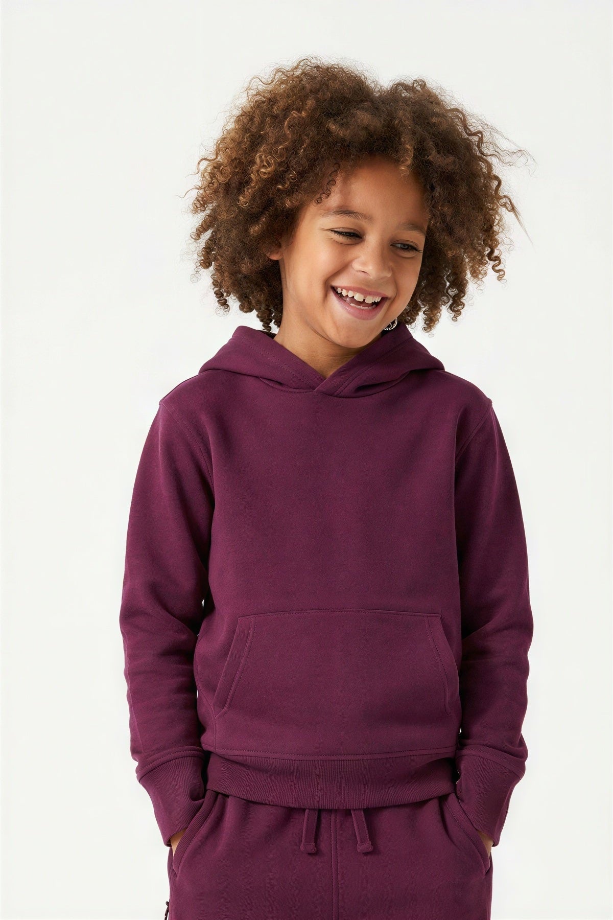 A smiling child with curly brown hair wearing a matching deep purple hoodie and sweatpants set, photographed against a clean white studio background. #burgundy