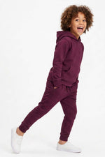 A child wearing a matching deep purple sweatshirt and jogger pants set, paired with white sneakers, posing against a solid white studio background. 