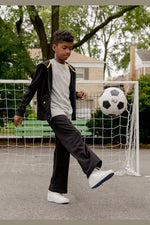 A young boy wearing a black zip-up hoodie with yellow shoulder accents, a white t-shirt, black sweatpants, and white sneakers kicks a soccer ball in front of a small soccer goal on an outdoor court. 