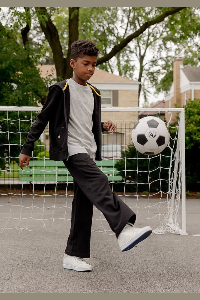 A young boy wearing a black zip-up hoodie with yellow shoulder accents, a white t-shirt, black sweatpants, and white sneakers kicks a soccer ball in front of a small soccer goal on an outdoor court. 