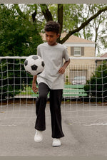 A child wearing a light gray oversized t-shirt, black wide-leg pants, and white sneakers is balancing a black and white soccer ball on their leg in front of a soccer net on an outdoor court. 