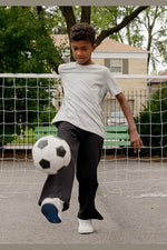 A young boy wearing a light gray t-shirt and dark gray sweatpants kicks a black and white soccer ball in an outdoor backyard setting with a net and trees in the background. 