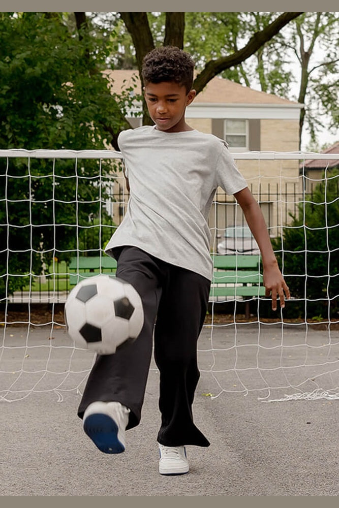 A young boy wearing a light gray t-shirt and dark gray sweatpants kicks a black and white soccer ball in an outdoor backyard setting with a net and trees in the background. 