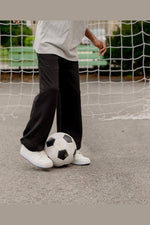 A person wearing loose black pants and white sneakers stands on a paved sports field near a soccer net, with a black-and-white soccer ball at their feet. 