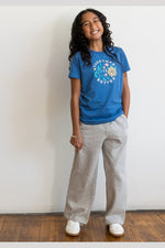 A young girl with long curly dark hair stands against a white wall wearing a blue graphic t-shirt, light gray wide-leg sweatpants, and white sneakers. 