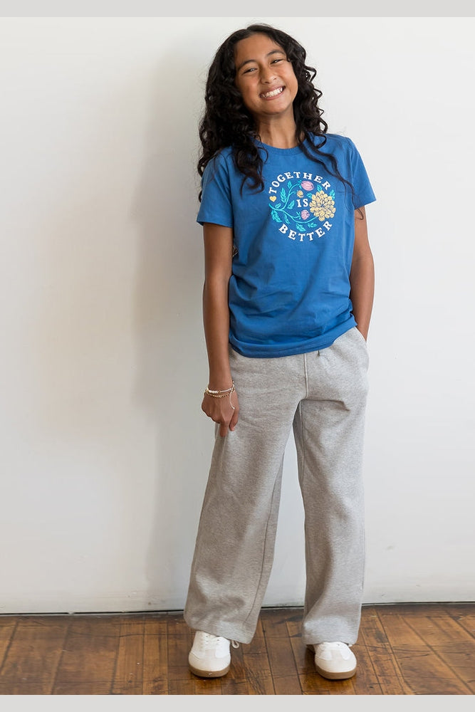 A young girl with long curly dark hair stands against a white wall wearing a blue graphic t-shirt, light gray wide-leg sweatpants, and white sneakers. 