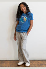 A young woman with long curly dark hair stands against a white wall wearing a blue graphic t-shirt, light gray wide-leg sweatpants, and white slip-on shoes. The t-shirt features a colorful circular graphic with the text 'BROOKLYN IS BETTER'. 