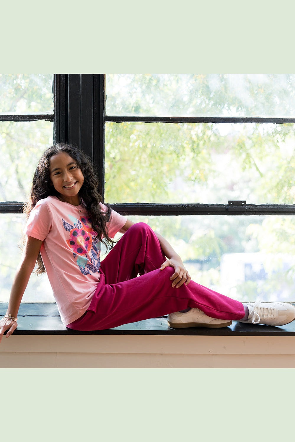 A young girl sitting on a windowsill wearing a light pink graphic t-shirt and bright magenta pink sweatpants, with white sneakers. The background shows a bright window with green foliage visible outside. 