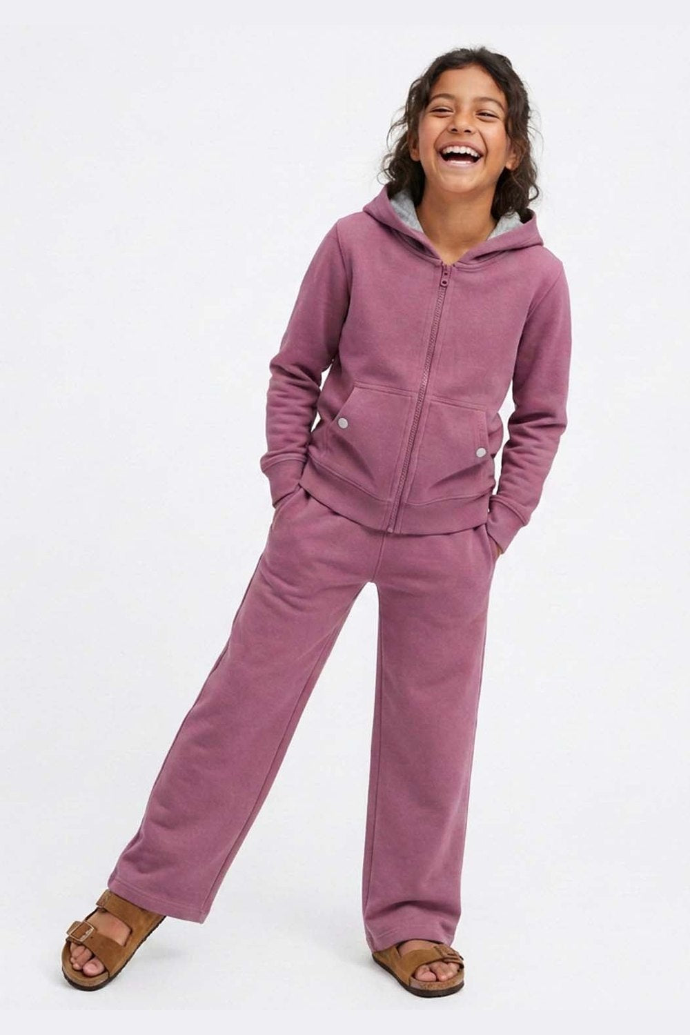 A young girl smiling and laughing while wearing a matching dusty rose purple tracksuit with a zip-up hoodie and wide-leg sweatpants, paired with brown slide sandals, against a plain white studio background. 