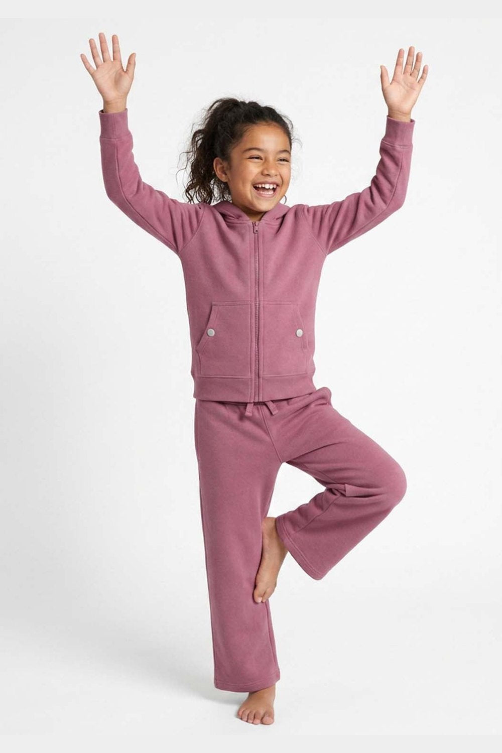 A young girl wearing a matching dusty rose fleece zip-up hoodie and sweatpants set, posing in a tree-balance pose against a clean white studio background. 