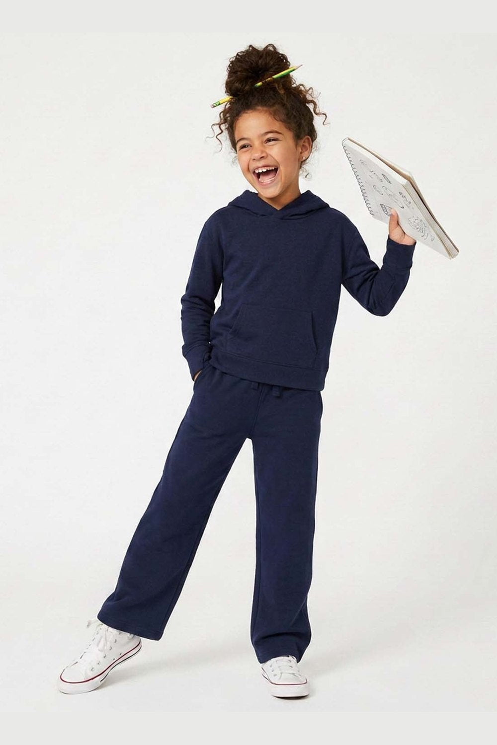 A young girl with curly hair wearing a matching navy blue sweatshirt and sweatpants set, smiling and laughing while holding a notebook against a plain white studio background. 
