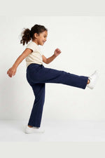 A young girl with curly hair is captured mid-motion, performing a high kick while wearing navy blue pants, a cream short-sleeve t-shirt, and white sneakers against a clean white studio background. 