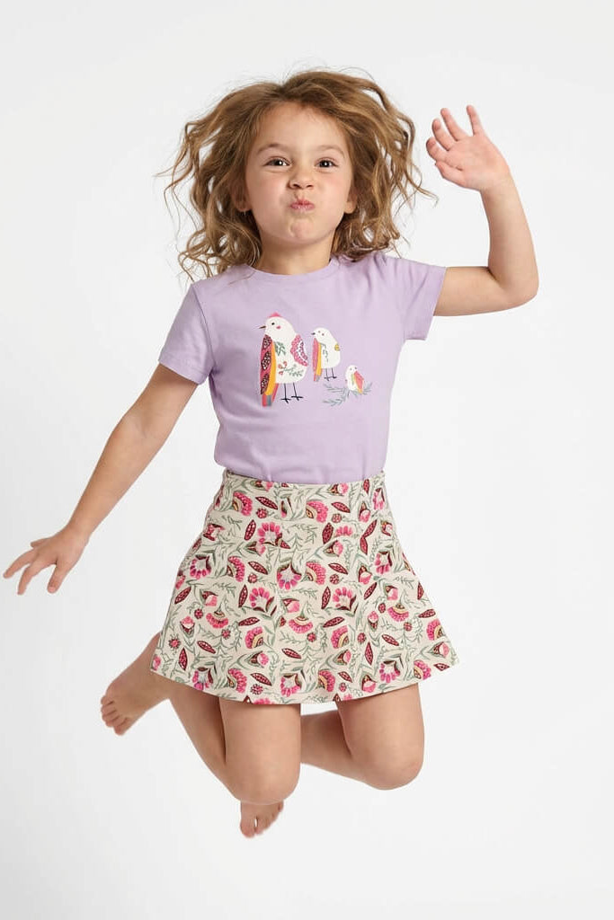 Young girl jumping. She is wearing a Mightly 100% organic cotton graphic tee with a colorful bird print and a floral skirt.  
