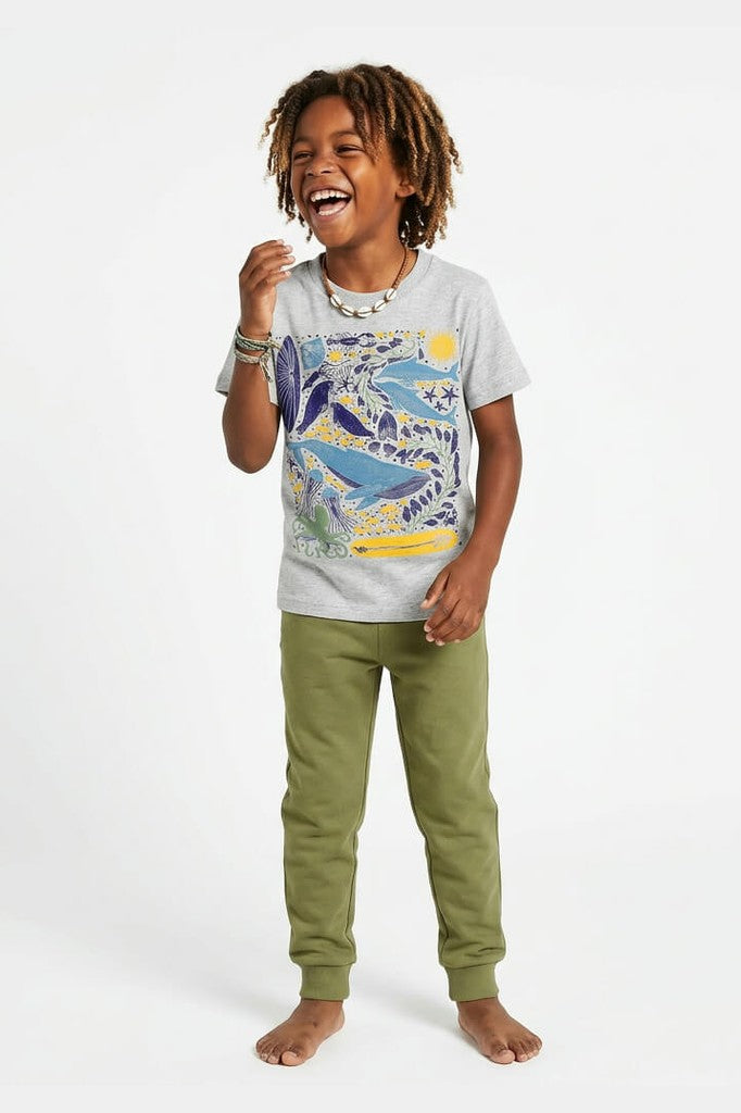Child wearing a gray t-shirt with a colorful whale design and green pants on a white background 