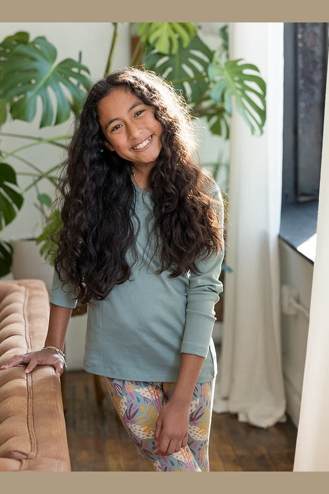 A smiling girl with long curly dark hair stands indoors wearing a sage green long-sleeve top and colorful patterned leggings, leaning against a tan sofa in a bright room with large green plants and a window. 