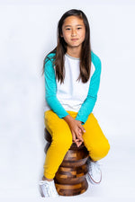 a girl sitting on a wooden stool wearing a teal and white long sleeve baseball tee and yellow pants