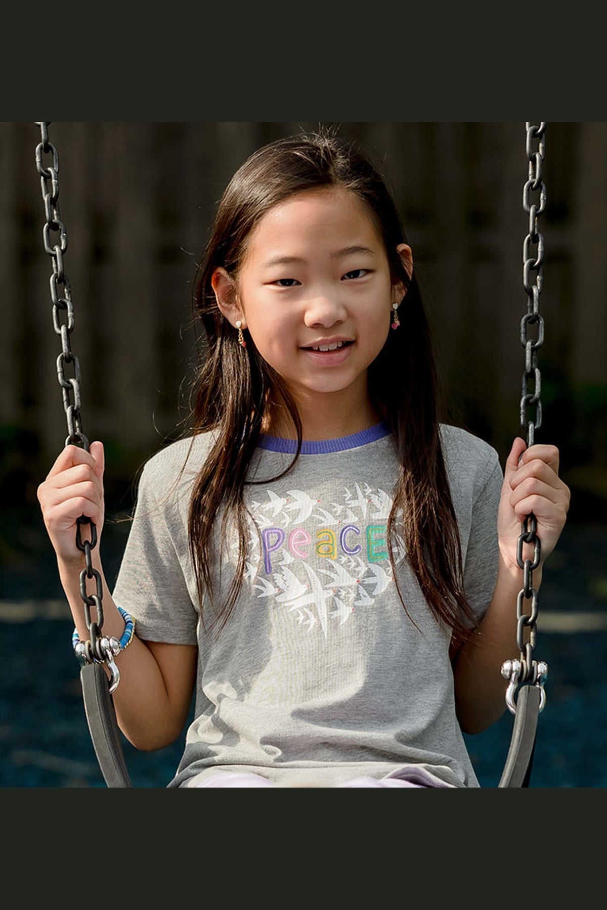 Young girl sitting on a swing wearing a t-shirt with 'Peace' printed on it.