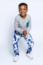 A young boy wearing a grey long sleeve tee with a striped pocket and tie dye sweatpants