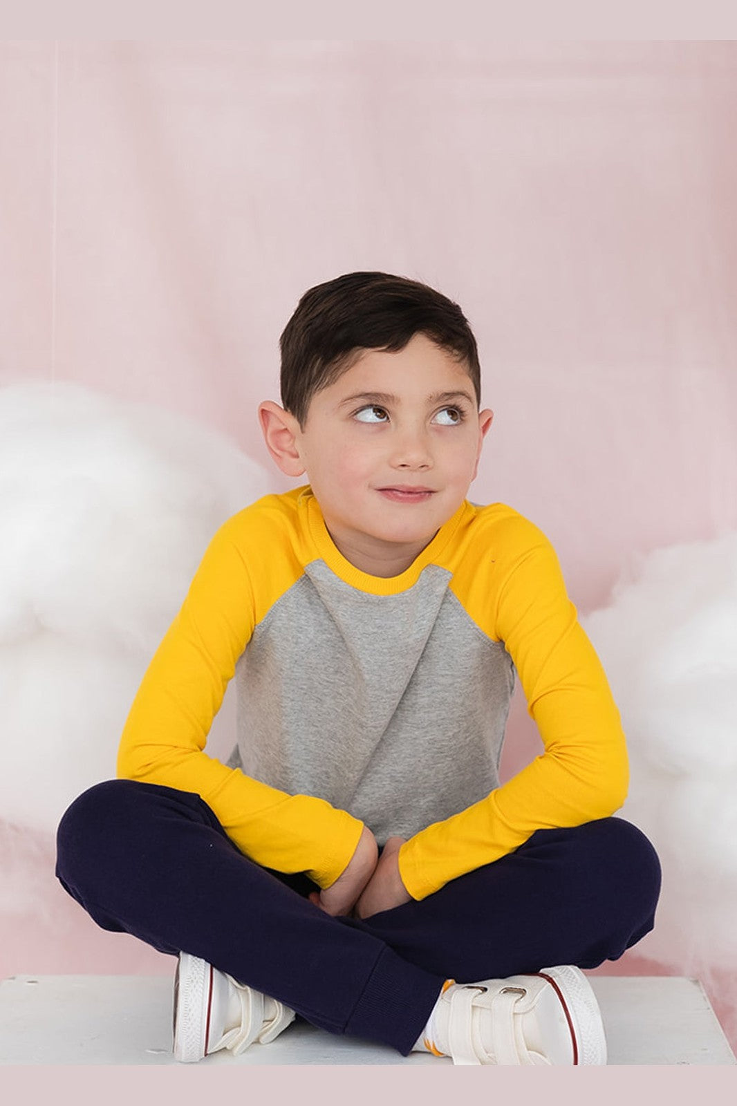 A boy sitting crossed legged on the floor wearing a gray and yellow long sleeve baseball tee