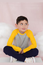 A boy sitting crossed legged on the floor wearing a gray and yellow long sleeve baseball tee