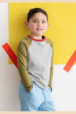 A young boy wearing a gray and green long sleeve baseball tee leaning against a colorful wall