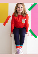 a young girl wearing a red long sleeve tee with a lightning bolt print pocket sitting on a bench against a colorful wall
