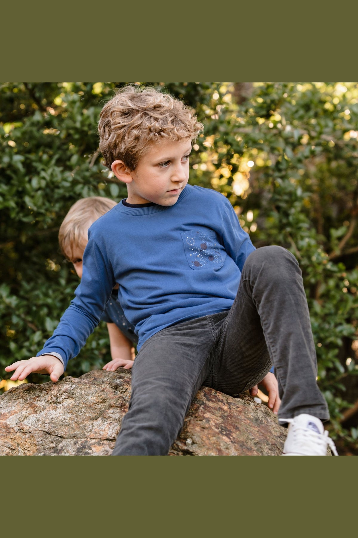 a young boy sitting on a rock wearing a blue long sleeve tee with a galaxy print on the pocket