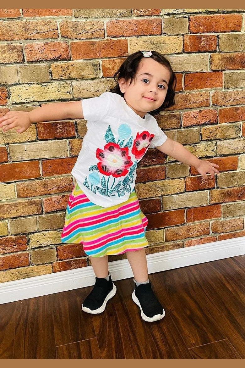 A young child wear a white shirt with a floral print and striped skirt against a brick wall