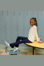 A young girl with braided hair sits on a yellow school table in front of blue-gray lockers. She is wearing a cream long-sleeve top, navy pants, white socks, and gray slip-on shoes. 