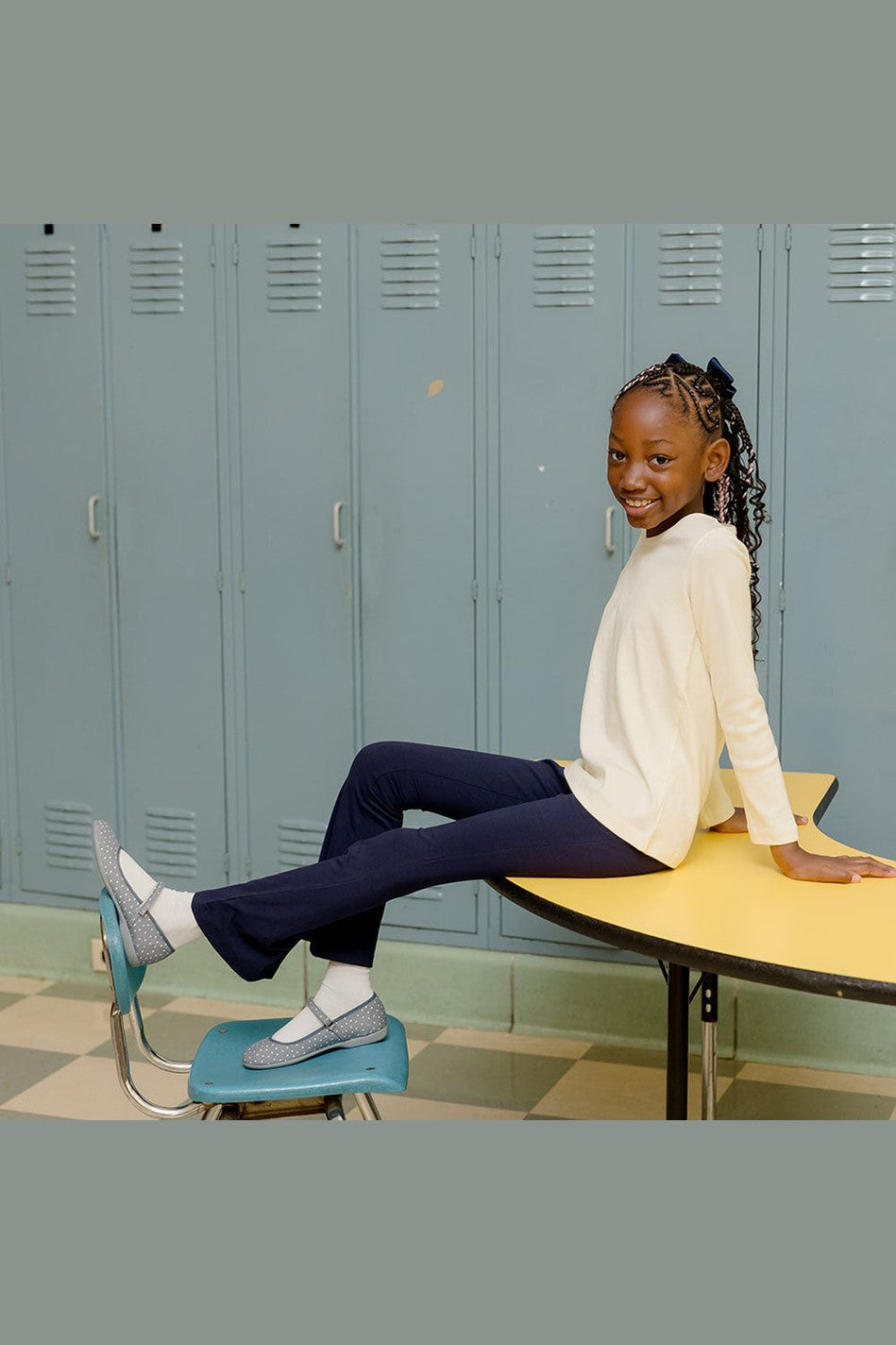 Girl wearing a long sleeve ivory shirt with navy blue flared leggings sitting on a desk in front of lockers