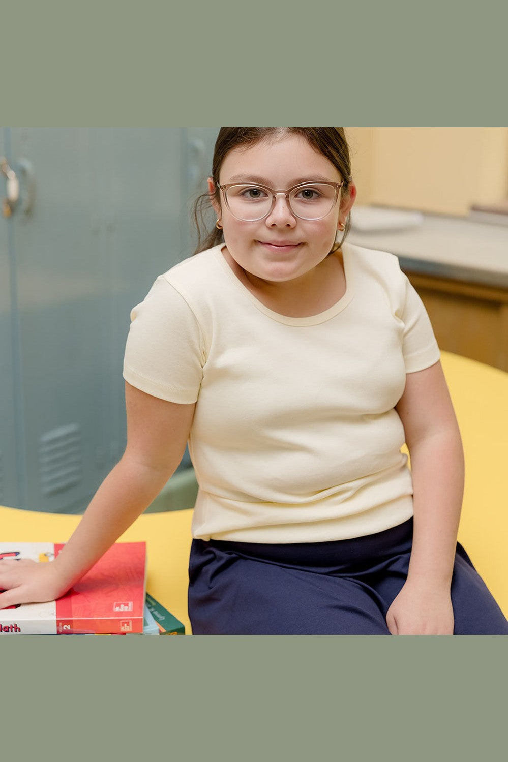 A young girl with glasses and brown hair tied back sits on a yellow desk in a classroom setting, wearing a cream-colored short-sleeve t-shirt and navy blue pants. 