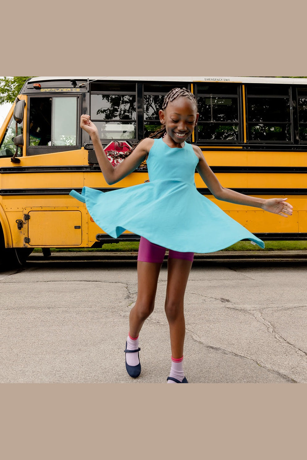 A girl twirling in a sleeveless turquoise dress and pink shorts with a school bus in the background